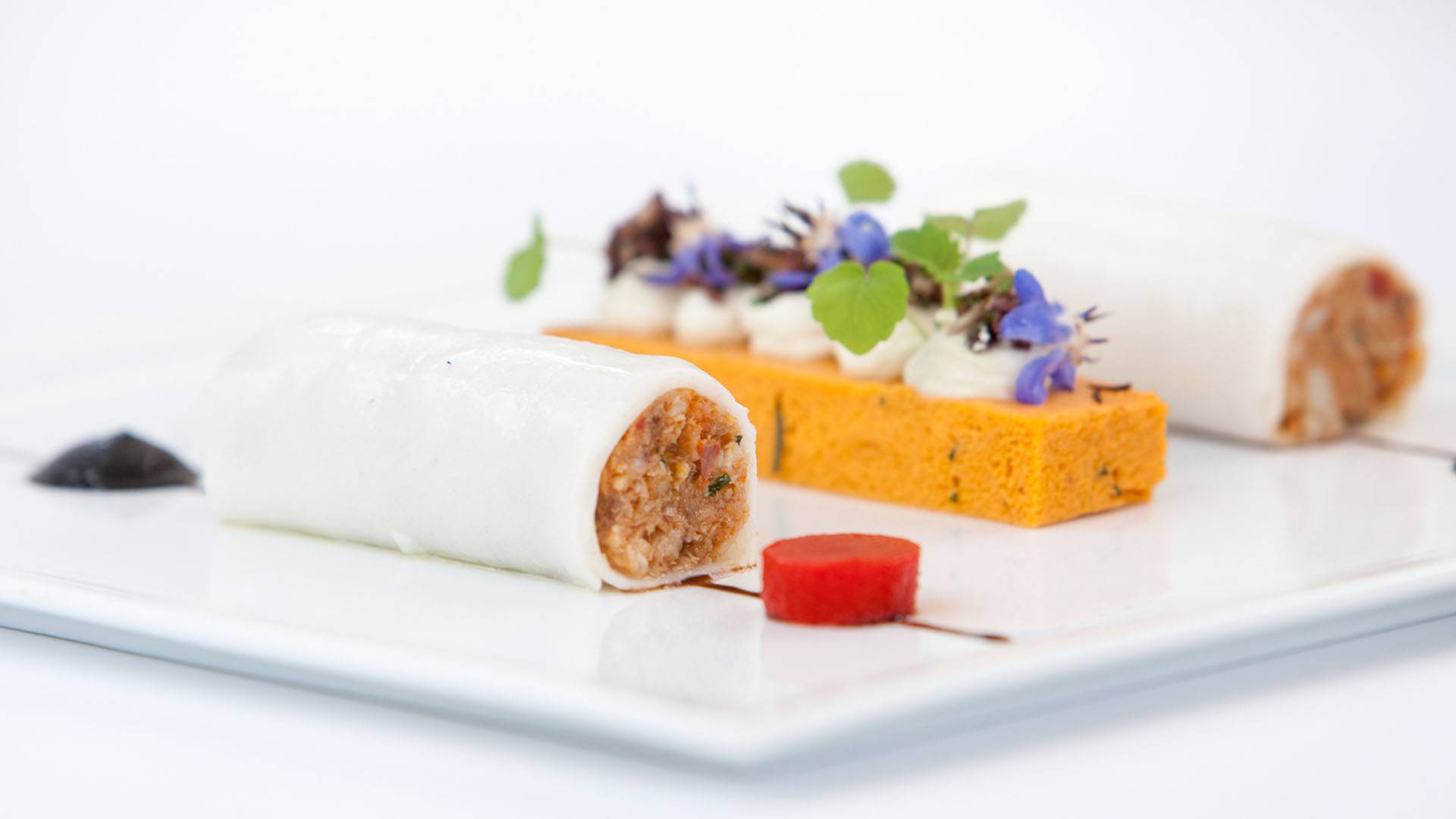 Two elegant food rolls with visible filling, orange sponge cake, herbs, edible flowers, and sauce dots on a white plate, evoking the conference's social dining occasions