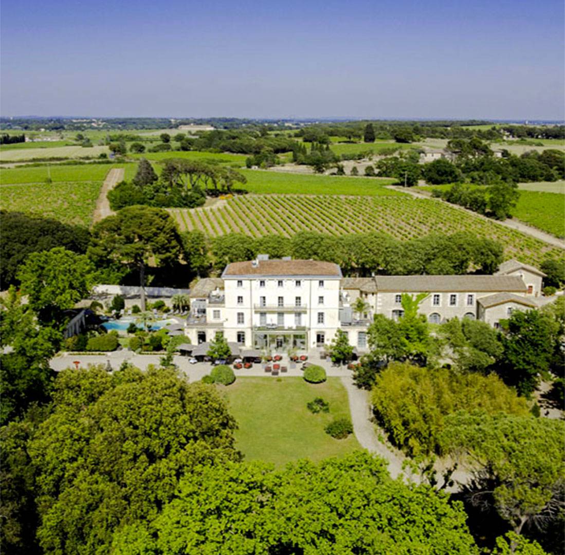 Aerial view of a large historic estate building surrounded by manicured gardens, vineyards, and countryside under a clear blue sky