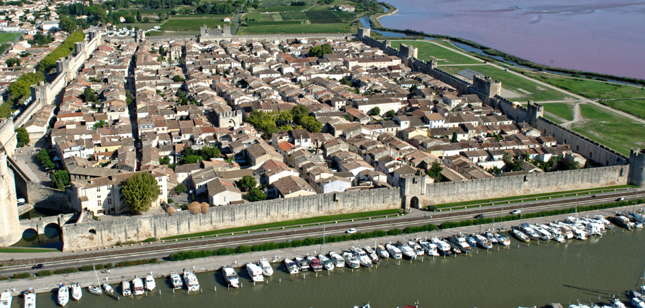 Aerial view of a walled historic town with dense rooftops, surrounded by fortified walls, a marina with boats, and nearby countryside under soft daylight