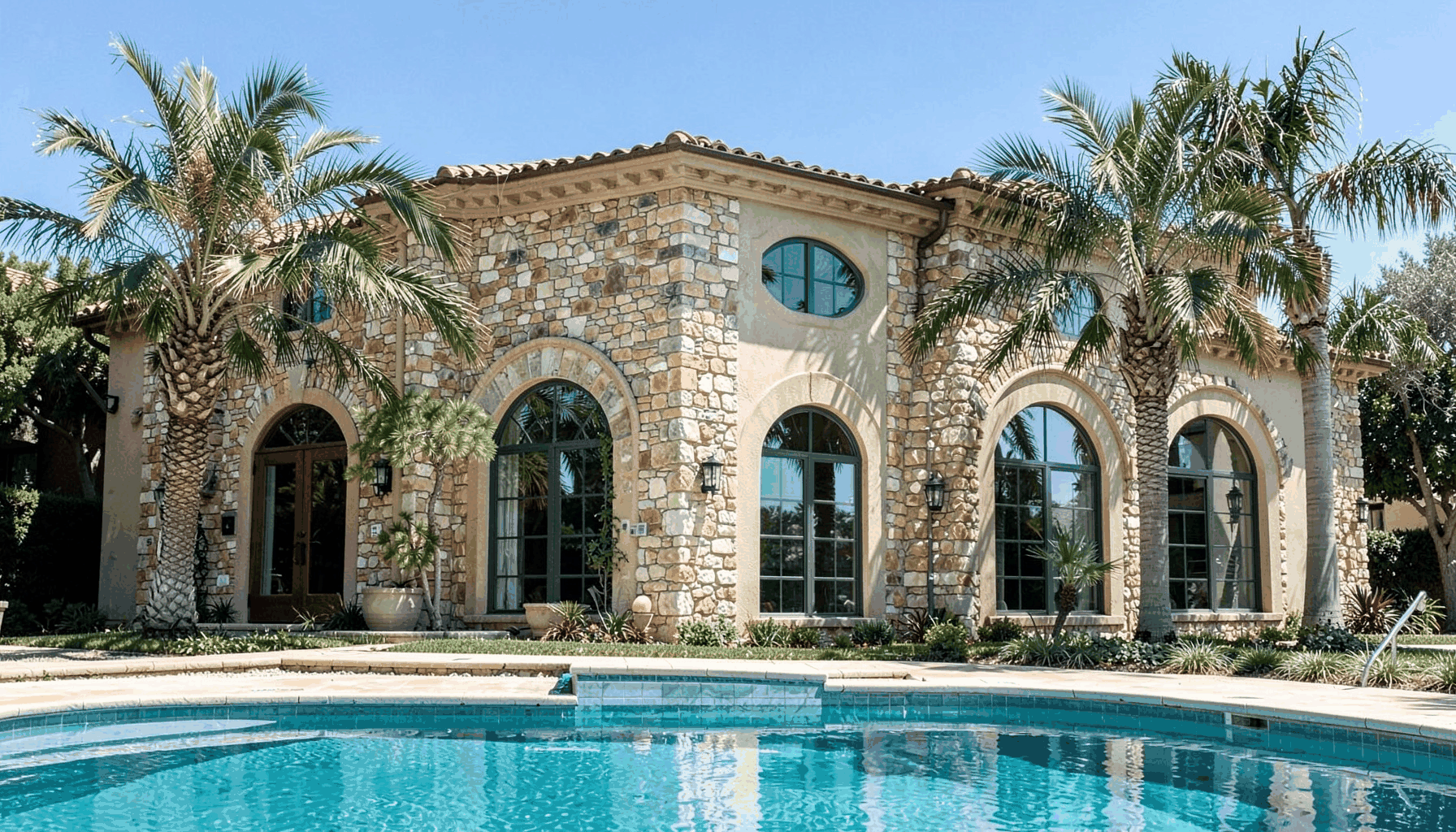 Stone and stucco building exterior with arched windows, palm trees, and a turquoise pool in the foreground under a clear sky